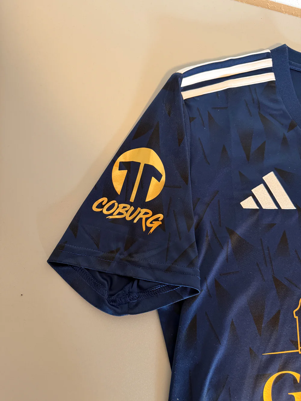 Goldener Sponsorenaufdruck Coburg auf dem Ärmel des dunkelblauen Jubiläumstrikots